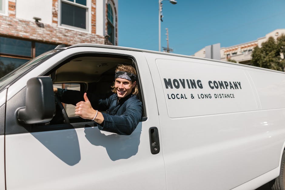 A young man with long, curly hair wearing a patterned bandana and a dark jacket is sitting in the driver's seat of a white moving van with the door open. He is smiling and giving a thumbs-up gesture w