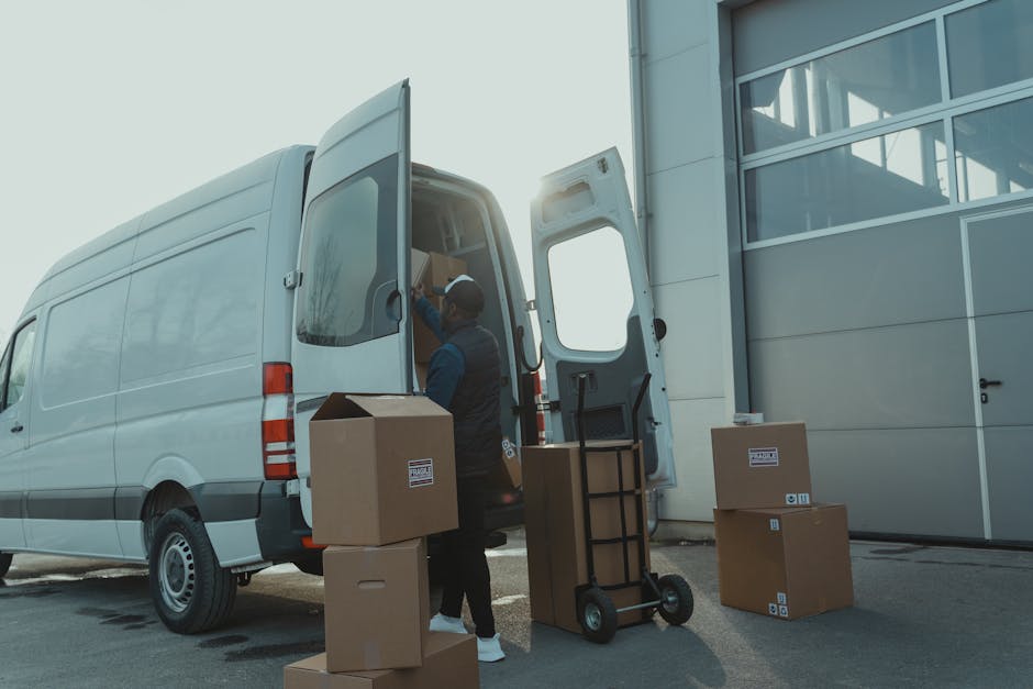 A white moving van parked on a city street with the logo 'MOVING COMPANY LOCAL & LONG DISTANCE' on its side. Inside the driver's side window, a woman wearing a dark jacket and gloves is seen managing