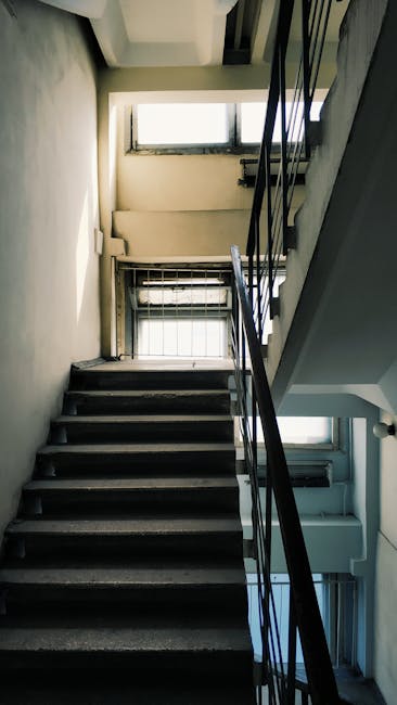 A vertical view of a narrow, indoor staircase in a UB6 tenement building in Perivale, showing dark wooden steps with a metal handrail on the right side. At the top of the staircase, there are two rectangular windows allowing natural light to illuminate the space, revealing a white wall and window frames. The lower window has a metal security grille, and the upper window appears foggy or frosted. The surrounding walls are painted light, neutral colours, with visible signs of wear. The context suggests a home relocation or furniture transport process, with no furniture or moving equipment visible in the image, but the setting is indicative of a building corridor used during moving or packing activities, as handled by Man with Van Perivale.