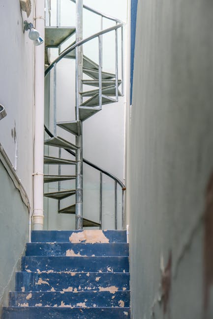A narrow, spiral metal staircase inside a residential property, featuring worn blue-painted wooden steps with peeling paint and visible scuff marks. The staircase has a black handrail attached to the wall on the right, and the surrounding walls are painted in light, neutral tones. Visible piping runs along the left side of the wall, and a small, grey, metal landings can be seen at the top of the staircase. The scene is lit by natural light coming from above, highlighting the compact space typical of tenement buildings in UB6 Perivale. This image depicts the challenging environment faced during a home relocation involving furniture transport through tight staircases, an area where specialist moving services like those offered by Man with Van Perivale are often required for safe and efficient removal and packing within such properties.