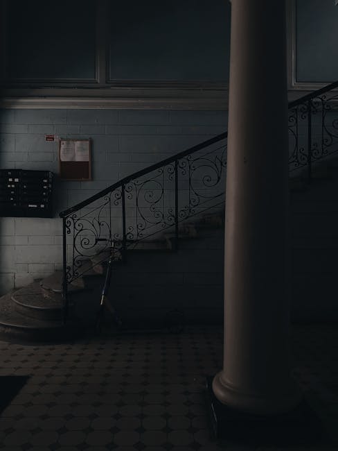 A vertical view of a narrow, indoor staircase in a UB6 tenement building in Perivale, showing dark wooden steps with a metal handrail on the right side. At the top of the staircase, there are two rectangular windows allowing natural light to illuminate the space, revealing a white wall and window frames. The lower window has a metal security grille, and the upper window appears foggy or frosted. The surrounding walls are painted light, neutral colours, with visible signs of wear. The context suggests a home relocation or furniture transport process, with no furniture or moving equipment visible in the image, but the setting is indicative of a building corridor used during moving or packing activities, as handled by Man with Van Perivale.