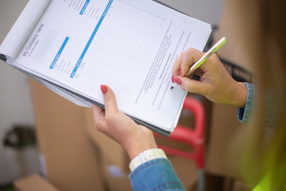 A person wearing a denim jacket and a white wristwatch is holding a clipboard with documents related to house removals, while using a yellow and green pen to make notes or sign the papers. The document on the clipboard has text and blue headers, indicating official paperwork, possibly permits or checklists associated with a home relocation or furniture transport. The individual is inside a property, perhaps in a hallway or near an entrance, with a blurred background showing cardboard boxes and a red trolley or dolly used for moving furniture, along with packaging materials like plastic wraps and packing paper. This scene illustrates the administrative and logistical aspects of a house removal completed by Man with Van Perivale, supporting efficient packing and moving processes, including permit arrangements if required by Ealing Council. The environment is well-lit, emphasizing the careful handling and organization involved in the removal process.