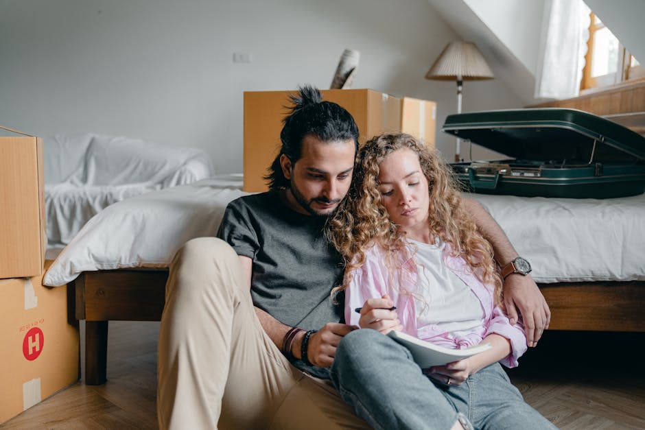An indoor scene showing a young man and woman sitting on the floor of a bedroom during a home relocation process. The man, with dark hair tied back, is dressed in a dark t-shirt and beige trousers, an