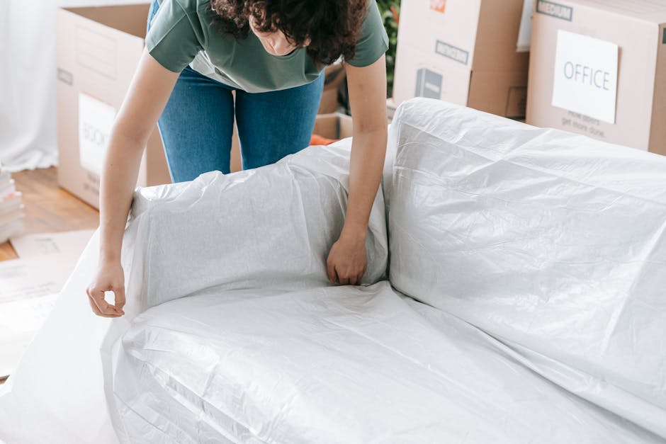 A person is seen preparing a large sofa for a home relocation inside a room, wrapping it with white packing paper or protective covering to safeguard the upholstery during furniture transport. The individual is bending over and carefully securing the covering around the sofa, which is positioned near a doorway with packed cardboard boxes and storage boxes labeled for office and other purposes visible in the background. The room has wooden flooring, and there are packing supplies nearby, indicating an organized move. This scene illustrates the packing and loading process involved in professional removals services, as provided by Man with Van Perivale, to facilitate efficient furniture transport during a house relocation or moving project.