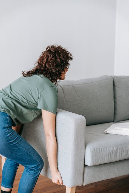 A person with curly brown hair wearing a light green shirt and blue jeans is inside a home, bending forward to lift a light grey fabric-covered sofa with wooden legs. The sofa is positioned against a white wall, and a second cushion or pillow is visible on the right side of the sofa. The room has wooden flooring, and the person appears to be involved in a packing and moving process, likely preparing the furniture for transport during a house removal. The scene reflects furniture transport and the loading process, consistent with home relocation activities supported by Man with Van Perivale.