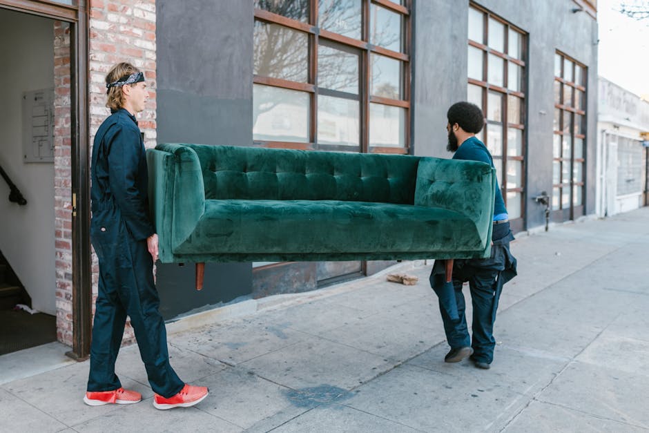 A person is seen preparing a large sofa for a home relocation inside a room, wrapping it with white packing paper or protective covering to safeguard the upholstery during furniture transport. The individual is bending over and carefully securing the covering around the sofa, which is positioned near a doorway with packed cardboard boxes and storage boxes labeled for office and other purposes visible in the background. The room has wooden flooring, and there are packing supplies nearby, indicating an organized move. This scene illustrates the packing and loading process involved in professional removals services, as provided by Man with Van Perivale, to facilitate efficient furniture transport during a house relocation or moving project.