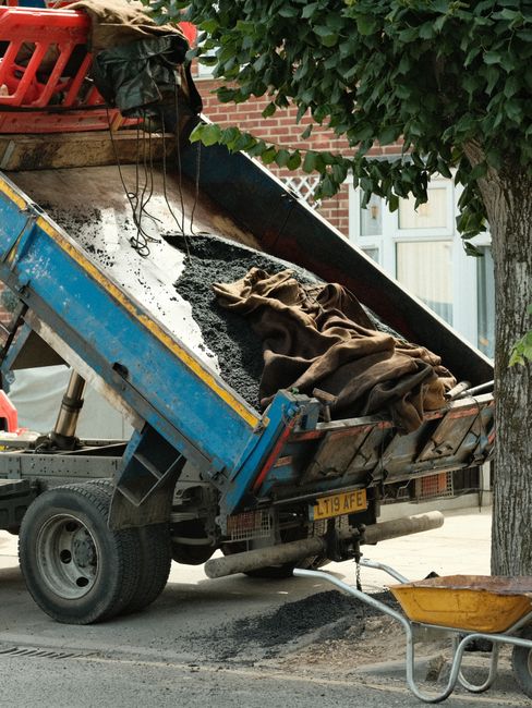 A blue tipper truck parked on a residential street during a house removal process, with its loading bed tilted upward for dumping a load of dark asphalt or similar material. The bed contains some brown fabric or soft padding and a partially covered pile of loose asphalt or gravel. The truck is adjacent to a curb, with a wheelbarrow positioned nearby, and is surrounded by trees and residential buildings with brick walls and glass windows, indicating a typical urban environment. The scene is illuminated by natural daylight, and the vehicle appears to be engaged in a building or paving-related task related to home relocation or infrastructure work, with the truck belonging to a removals or construction service.
