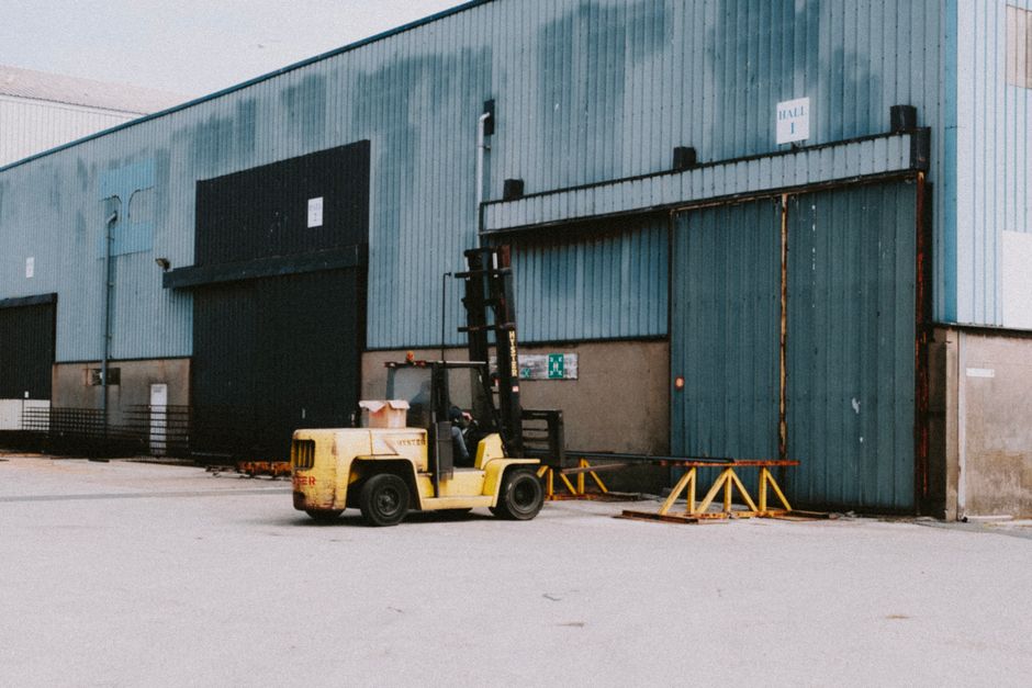 A yellow forklift truck with black accents is parked outside a large, industrial warehouse building with a corrugated metal exterior painted in shades of blue and grey. The forklift is positioned on a paved surface and appears to be in the process of loading or unloading goods. In front of the forklift, there are orange and yellow safety barriers and a small collection of cardboard boxes and protective packaging materials, likely used for a house relocation or furniture transport. The warehouse features several large sliding doors, some of which are partially open, revealing the interior space, while others are closed. The scene is set during daylight hours, with natural light illuminating the area, and the environment suggests it is part of a professional removals or moving service operation, such as those provided by Man with Van Perivale, specializing in packing, loading, and transport logistics for home removals. The overall setting emphasizes the logistics involved in furniture movement and building access for a home relocation process.