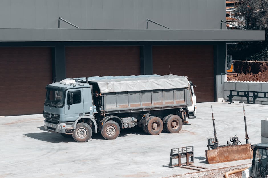 A blue tipper truck parked on a residential street during a house removal process, with its loading bed tilted upward for dumping a load of dark asphalt or similar material. The bed contains some brown fabric or soft padding and a partially covered pile of loose asphalt or gravel. The truck is adjacent to a curb, with a wheelbarrow positioned nearby, and is surrounded by trees and residential buildings with brick walls and glass windows, indicating a typical urban environment. The scene is illuminated by natural daylight, and the vehicle appears to be engaged in a building or paving-related task related to home relocation or infrastructure work, with the truck belonging to a removals or construction service.