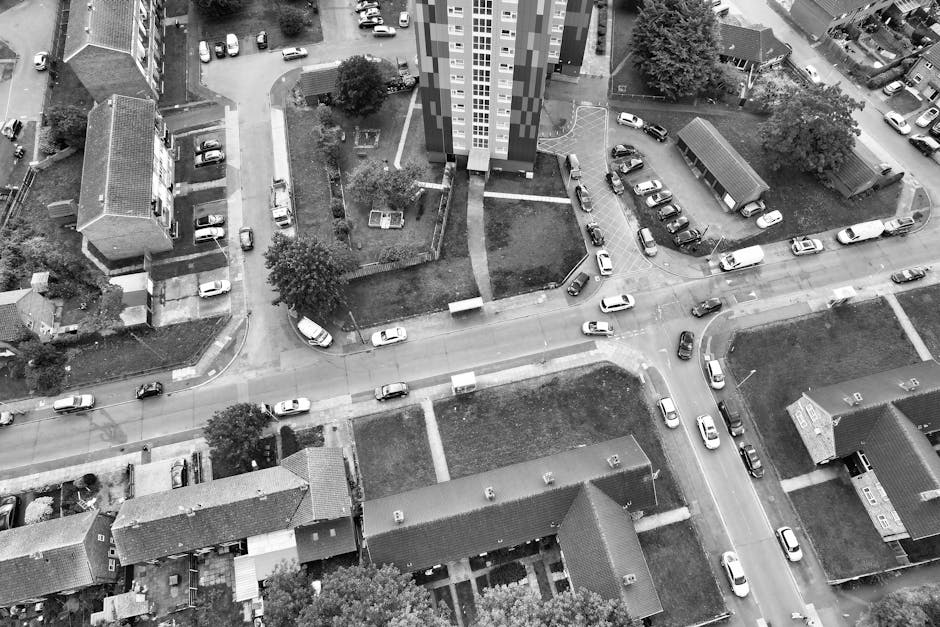 An aerial black-and-white photograph showing a suburban residential area with several multi-storey apartment buildings, individual houses with pitched roofs, and a network of roads with parked and moving cars. In the foreground, there are rows of narrow row houses with small front gardens and driveways. The streets intersect, and some vehicles are paused or moving, indicating typical traffic flow. There are patches of greenery, including trees and grass areas, scattered throughout the scene. The image also captures the loading process for house removals, with furniture, cardboard boxes, and packing materials visible in one of the driveways, being prepared for transport by a professional moving company like Man with Van Perivale. The environment appears well-lit with natural daylight, and the overall scene reflects the logistical aspects of home relocation, involving furniture transport and packing in a residential setting.