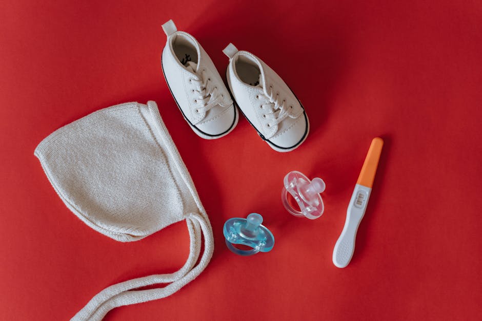 A flat lay arrangement on a red surface showing a pair of small white canvas sneakers with black trim, a white cloth baby hat with ties, two pacifiers (one blue and one pink), and an orange and white digital thermometer. The items are positioned at the center of the image, with the sneakers at the top, the hat to the left, the pacifiers below the hat, and the thermometer to the right. This arrangement depicts items that might be packed or prepared during a home relocation or packing process, such as for moving to a new residence, consistent with house removals services offered by Man with Van Perivale.