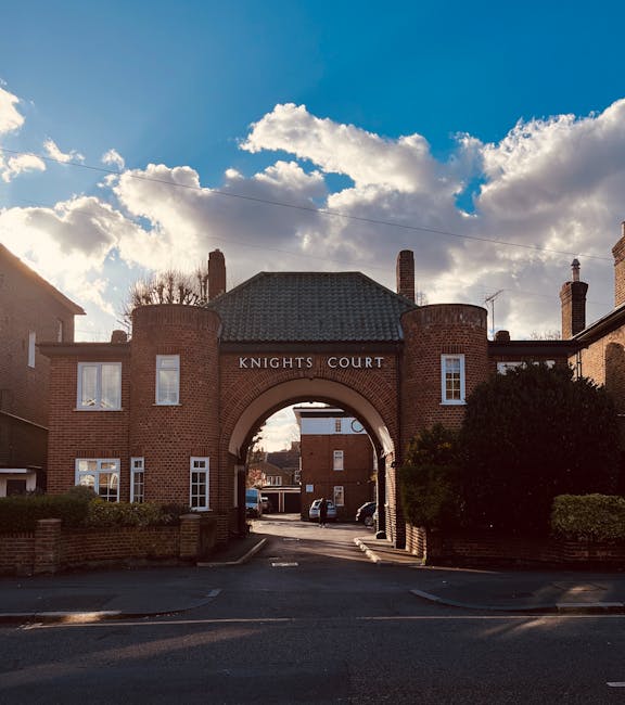 Image of the entrance to Knights Court, a residential housing complex constructed from red brick, featuring a large central archway and two symmetrical towers on either side. The archway leads to an internal street where parked cars are visible. Above the arch, the name 'KNIGHTS COURT' is displayed in white lettering. The sky overhead is partly cloudy, with sunlight illuminating the structure and casting shadows on the surrounding greenery and pavement. Trees and bushes border the entrance on both sides, and the scene captures a quiet moment in an urban setting. The photograph provides a clear view of the building’s architecture, suitable for illustrating aspects of property access or house removals related to home relocation services provided by companies like Man with Van Perivale.