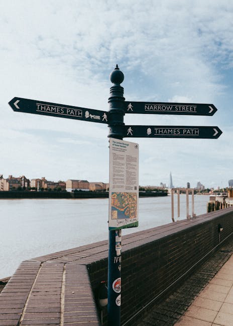 A signpost situated on the pavement beside the Thames River in Perivale's narrow streets area, indicating directions to Thames Path and Narrow Street, with a map and instructions attached to the pole. In the background, the river is visible with some modern buildings along the opposite bank, and a partly cloudy sky overhead. This setting illustrates an outdoor location relevant for planning house removals and home relocation logistics. During a moving process, such signage can assist in understanding local navigation routes for efficient furniture transport through urban, narrow streets, and along riverside routes, with the signpost serving as a point of reference for directional orientation in moving operations, which may be arranged by companies like Man with Van Perivale.