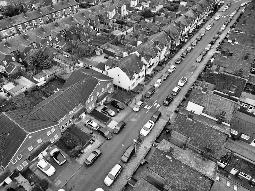 A black and white aerial photograph of a residential street with closely packed terraced houses and detached homes in a suburban neighbourhood. Cars are parked along the sides of the narrow pavement and in designated parking bays fronting each property. The street includes a single lane in each direction, marked with white lines and directional arrows. In the foreground, a move is taking place with a man from Man with Van Perivale overseeing the loading process, where a large cardboard box and wrapped furniture, including a wooden cabinet, are being carefully carried onto a medium-sized van parked on the street. The vans are positioned parallel to the pavement with some partially loaded, and are part of a home relocation or furniture transport operation involving packing and moving household items. The environment is well-lit, with soft natural light illuminating the scene, and the surrounding houses have tiled roofs, chimneys, small gardens, and fence boundaries typical of the area, highlighting the urban context for a professional removal service.