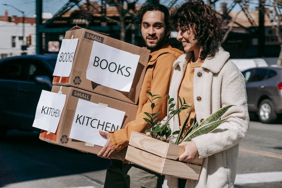 Two individuals are outdoors on a paved street, engaged in house relocation activities. The man on the left is holding two cardboard boxes, labeled 'BOOKS' and 'KITCHEN,' which are taped shut and appear to contain packed belongings. The woman on the right is carrying a wooden crate filled with potted plants and leafy foliage, suggesting they are in the process of packing or unloading household items. Both are dressed in casual, neutral-toned clothing suitable for moving tasks, and they are smiling and talking. In the background, a moving van or vehicle with an open door is visible, along with parked cars and residential buildings, indicating a neighbourhood setting. The scene is lit by natural daylight, with shadows cast on the pavement, and the overall environment reflects a typical house removal operation involving packing, furniture transport, and logistical planning, consistent with services offered by Man with Van Perivale in house removals and relocations.