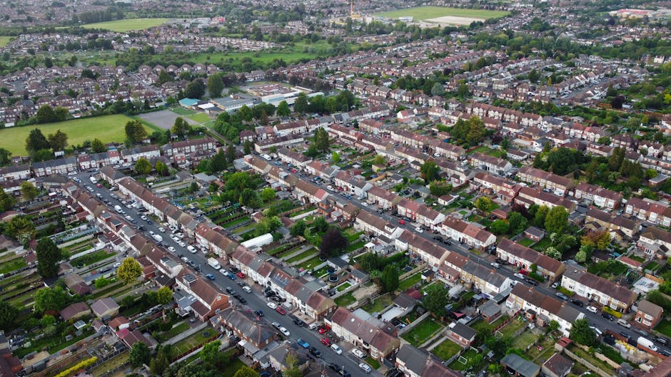 An aerial view of a suburban residential area showing rows of terraced houses with small front gardens and driveways, lined along narrow streets with parked cars on both sides. The houses have a variety of roof types, including tiled and flat roofs, with some featuring chimneys, and are mostly brick-built. Green trees and lawns are visible interspersed throughout the neighbourhood, with larger open green spaces and sports fields in the distance. The scene includes a few larger buildings and playgrounds amongst the houses, under an overcast sky with diffuse natural lighting. This image captures the layout typical of home relocation and furniture transport contexts, highlighting the densely packed housing environment within a community that may experience restrictions on moving days, as referenced in the PERIVALE article. Man with Van Perivale occasionally assists with packing, lifting, and transporting furniture during such residential moves.